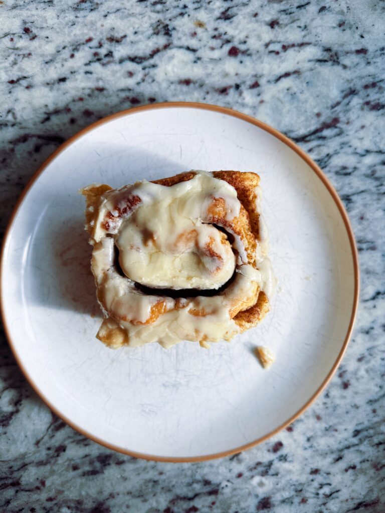 A delicious gluten-free cinnamon roll topped with cream cheese frosting on a white plate, set against a textured granite countertop.