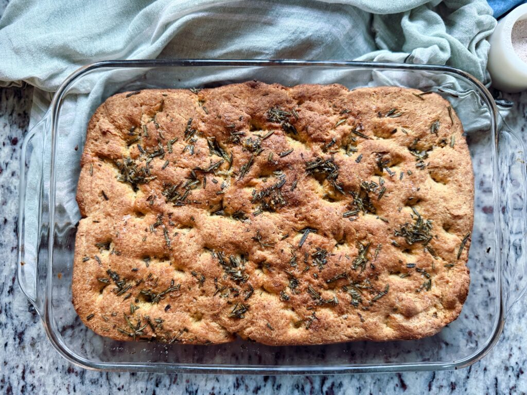A freshly baked focaccia bread topped with herbs, presented in a glass baking dish on a granite countertop.