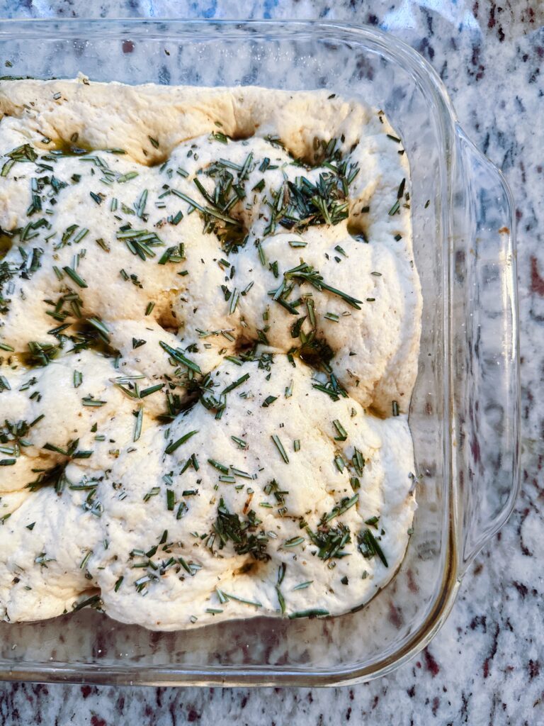 A close-up view of uncooked gluten-free focaccia dough topped with fresh rosemary in a glass baking dish.