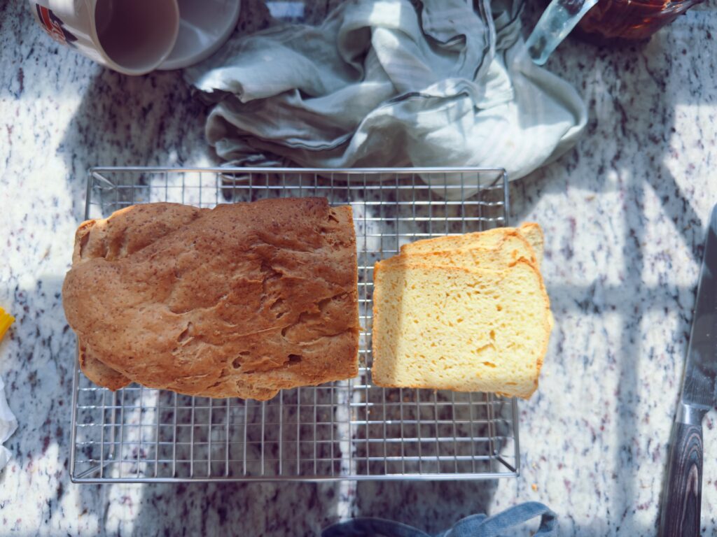 A loaf of freshly baked bread on a cooling rack, with one slice cut and placed beside it, set on a granite countertop with kitchen items in the background.