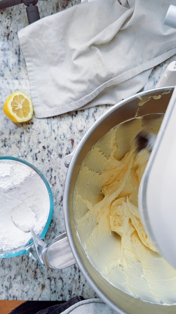 A bowl of creamy lemon buttercream frosting is shown being mixed in a stand mixer, with a portion of powdered sugar in a bowl nearby and a lemon slice on a marble countertop.
