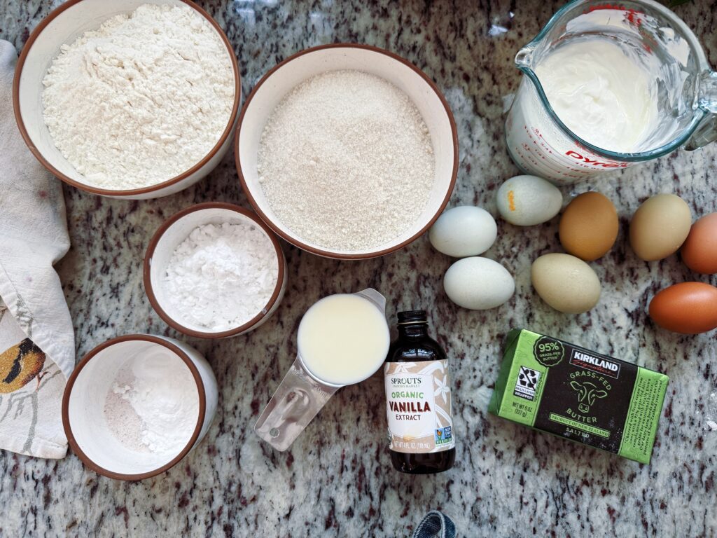 Ingredients for baking a gluten-free blueberry cake laid out on a marble countertop, including bowls of various flours, sugar, eggs, milk, butter, and vanilla extract.