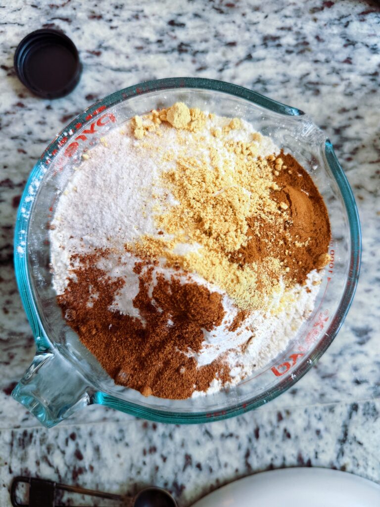 Close-up of a measuring bowl filled with various dry ingredients, including gluten-free flour, spices like cinnamon and ginger, and baking soda, set on a granite countertop.