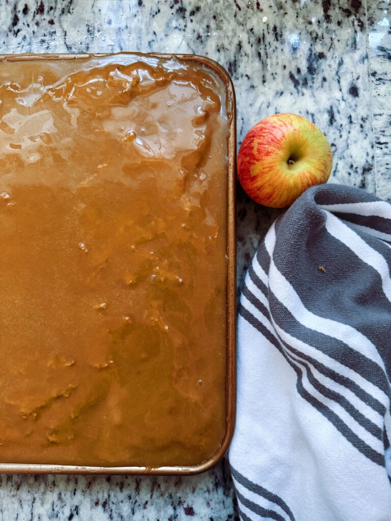 A gluten-free apple spice sheet cake topped with a brown sugar frosting, placed on a speckled countertop alongside a fresh apple and a striped dish towel.