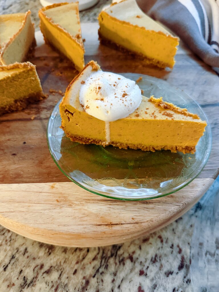 A slice of gluten-free pumpkin cheesecake topped with whipped cream and a sprinkle of cinnamon on a glass plate, with additional slices in the background.