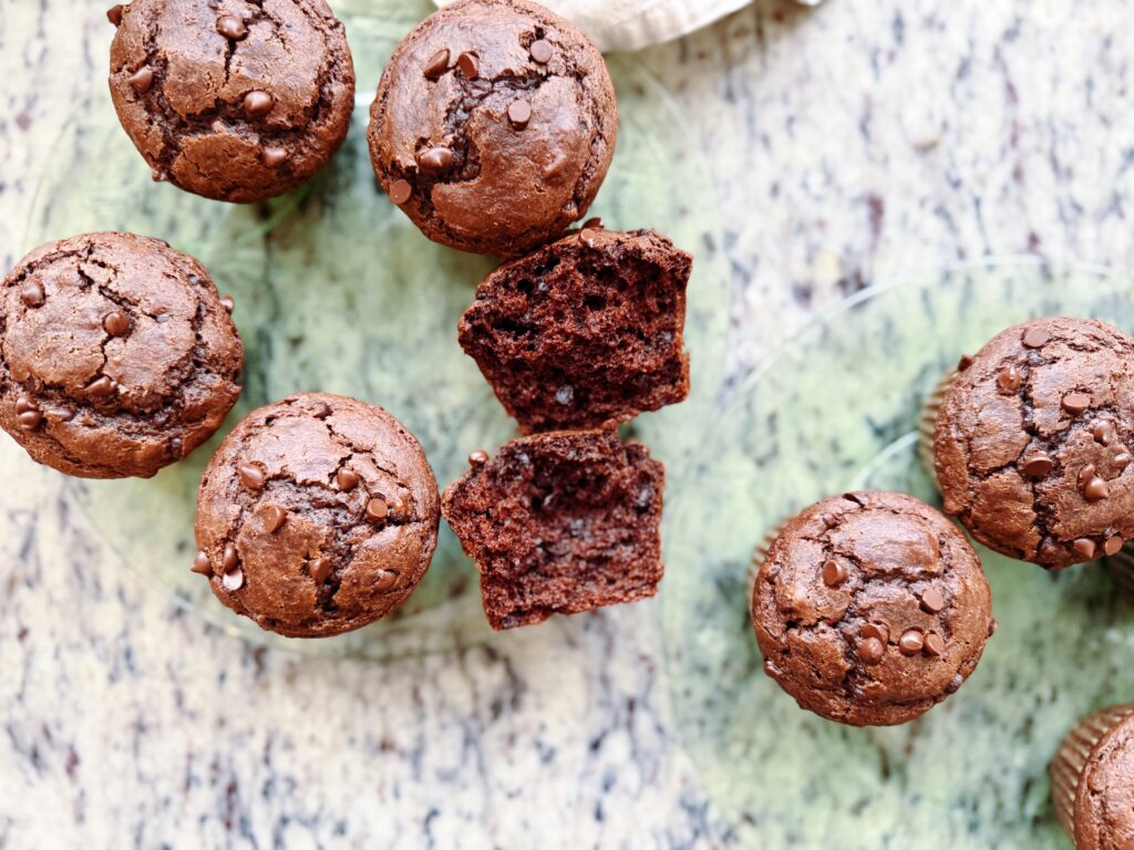 A top-down view of fresh chocolate muffins displayed on a glass plate, with one muffin cut in half to show its moist interior and chocolate chips.