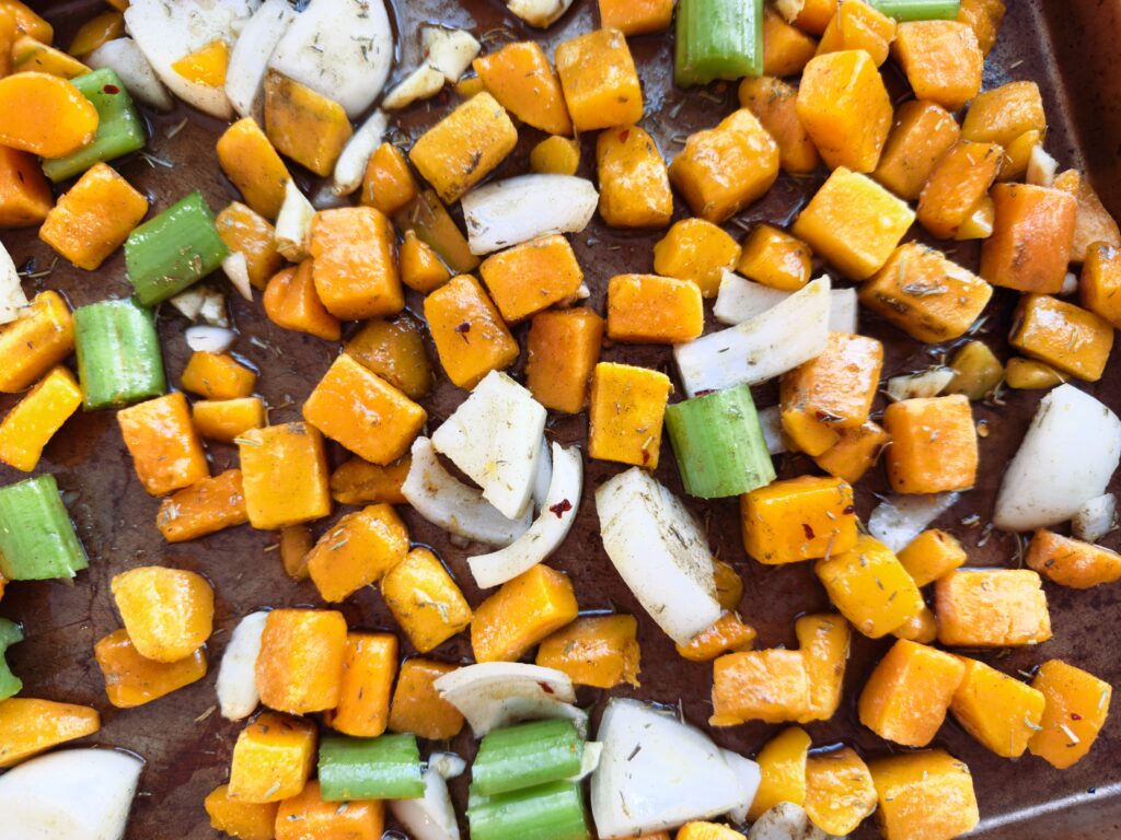 Chopped butternut squash, carrots, celery, and onions drizzled with olive oil and herbs on a baking sheet, ready for roasting.