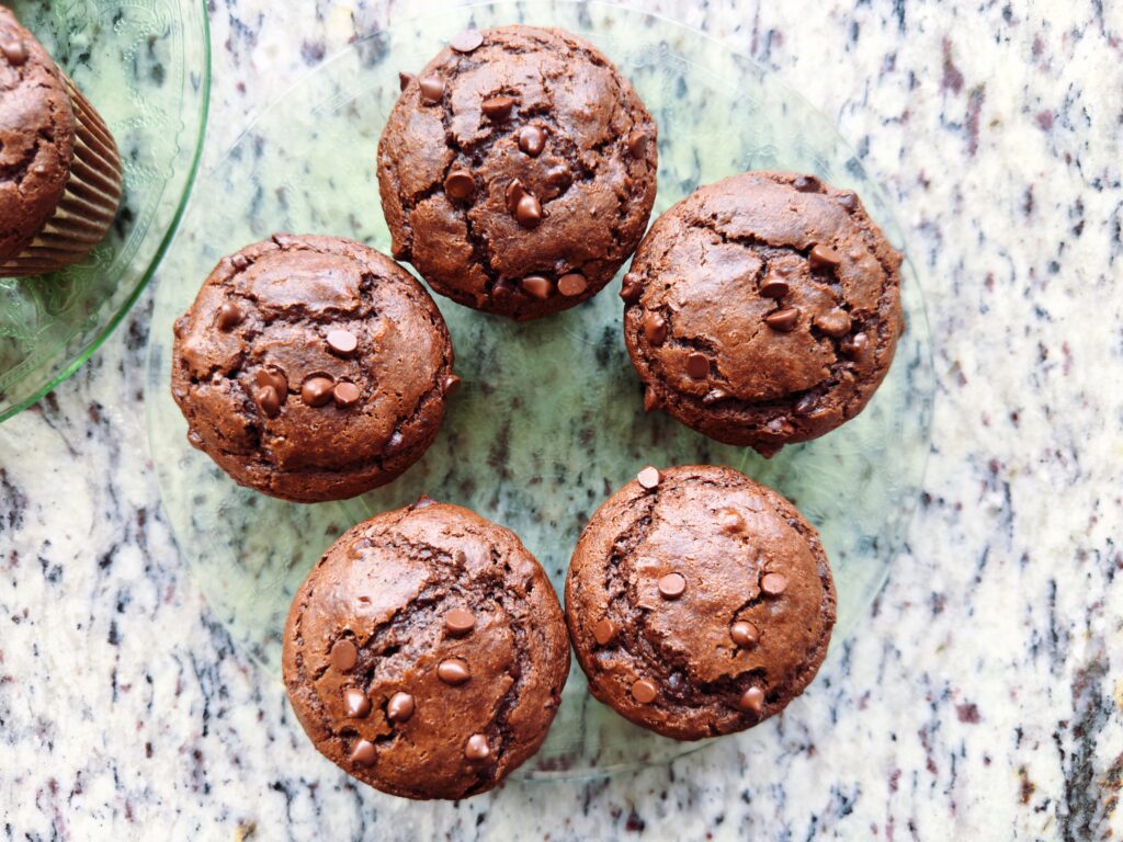 A top-down view of five chocolate muffins arranged on a glass plate, showcasing chocolate chips on the surface.
