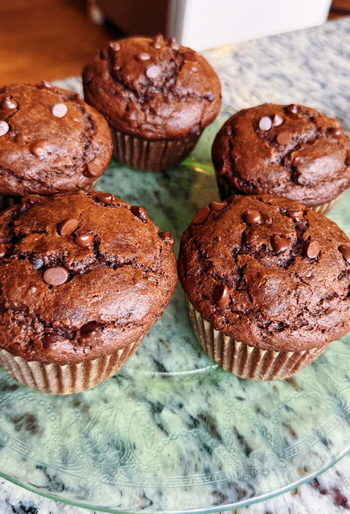 A close-up of five chocolate muffins with chocolate chips on top, arranged on a decorative plate against a marble background.