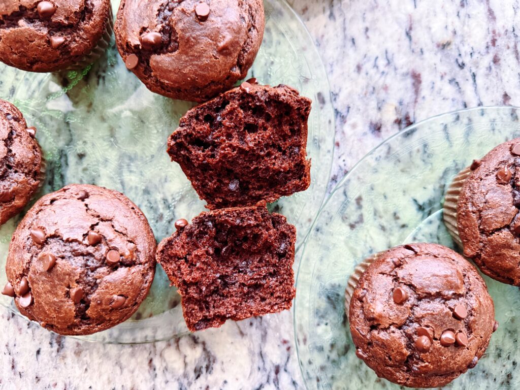 A close-up of chocolate muffins on a glass plate, with one muffin cut in half to reveal its fluffy interior and chocolate chips.