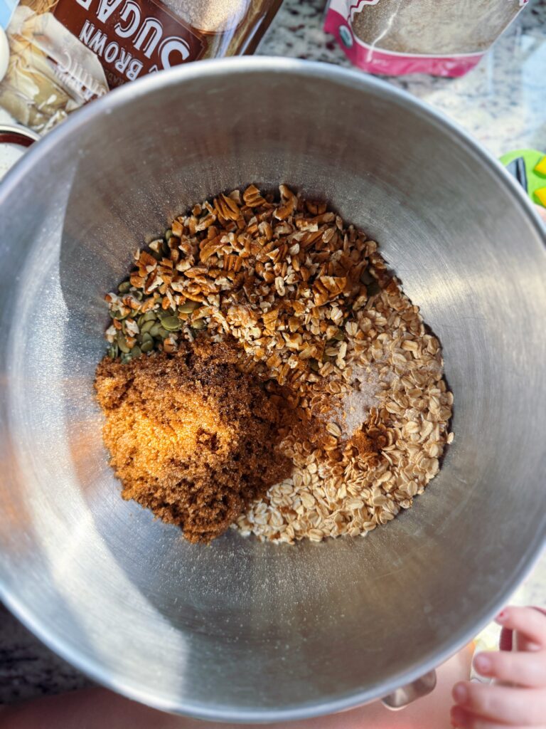 A mixing bowl containing a variety of dry ingredients including oats, chopped nuts, seeds, and brown sugar, with sunlight illuminating the contents.