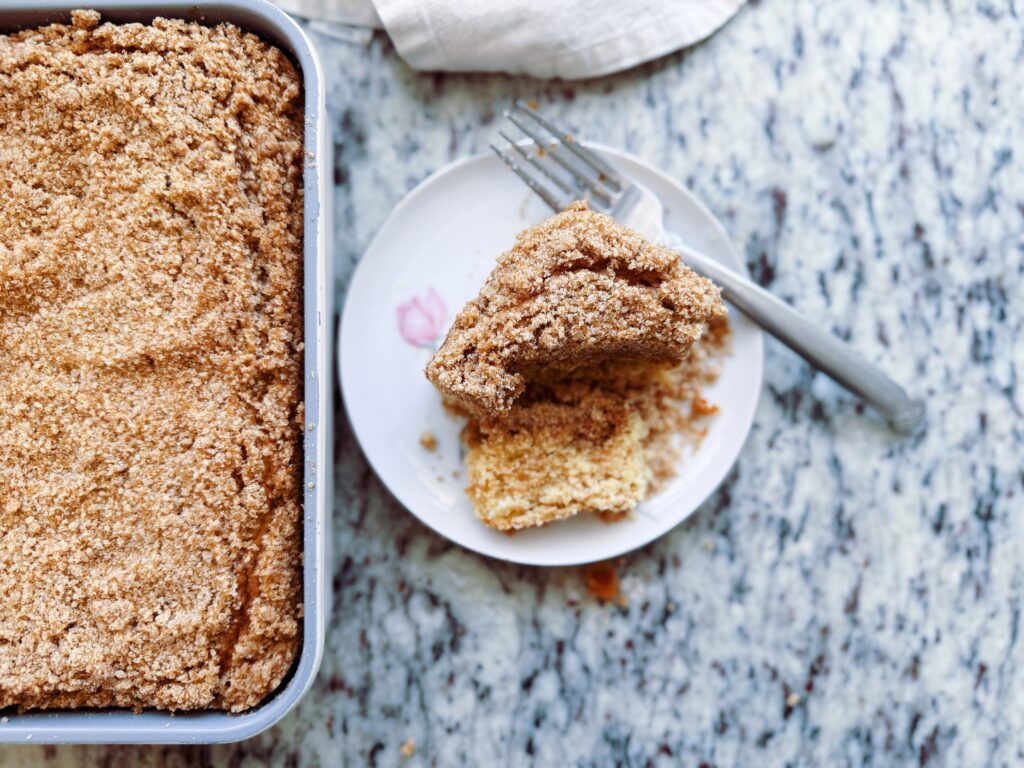 A close-up of a crumbly dessert in a baking dish next to a plate with a piece of the dessert, placed on a marble surface.
