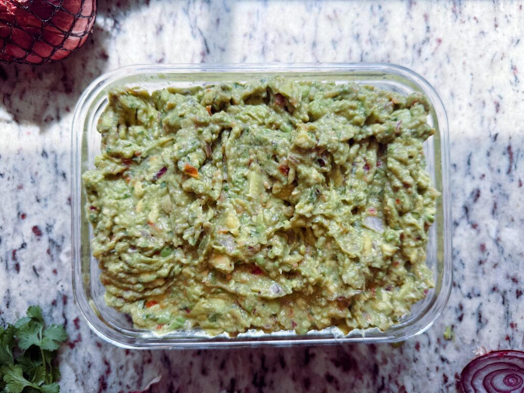 A glass container filled with creamy guacamole on a marble countertop, garnished with chopped onions and cilantro.