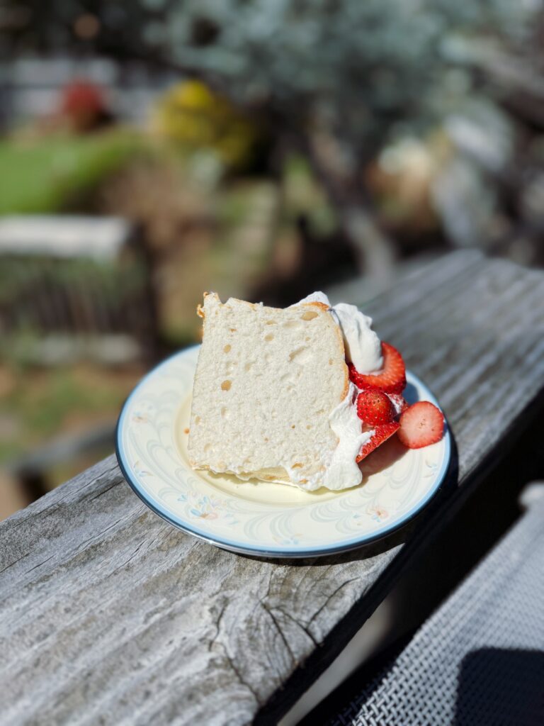 A slice of cake topped with whipped cream and strawberries on a decorative plate, resting on a wooden surface, with a blurred garden background.
