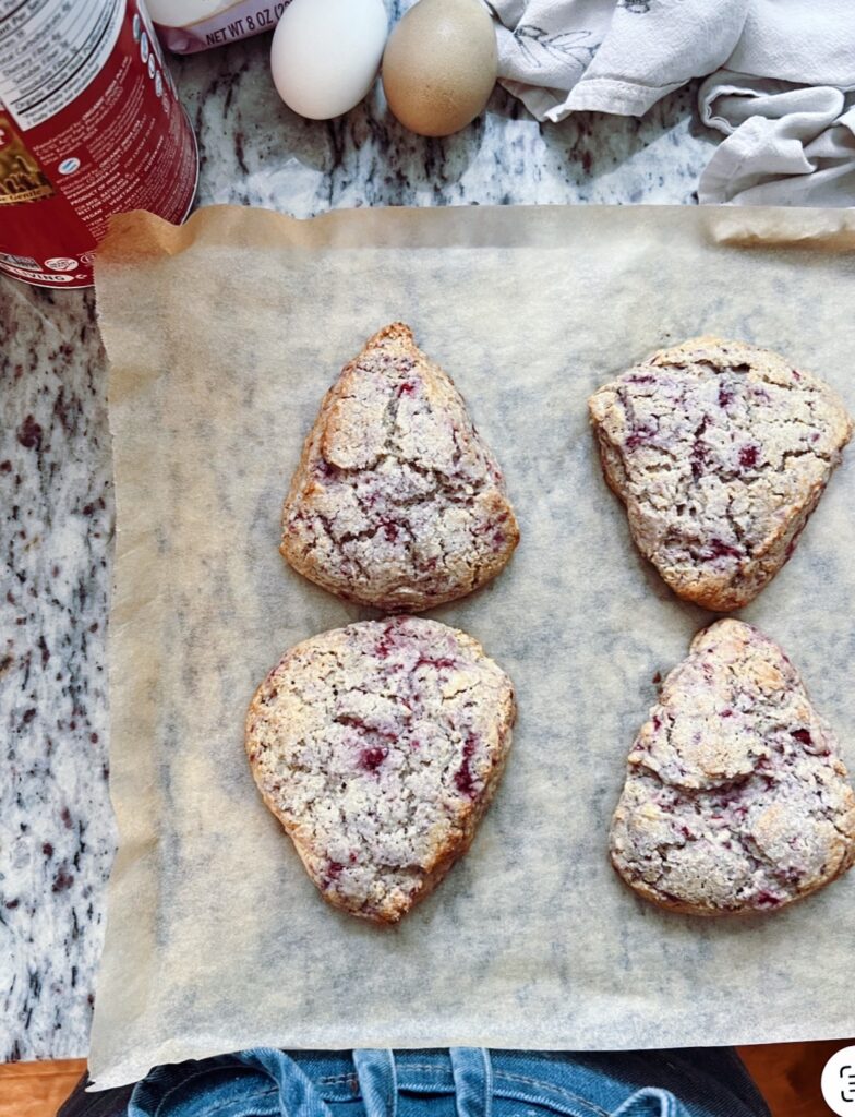 Four freshly baked scones on parchment paper, with a few ingredients like eggs in the background.