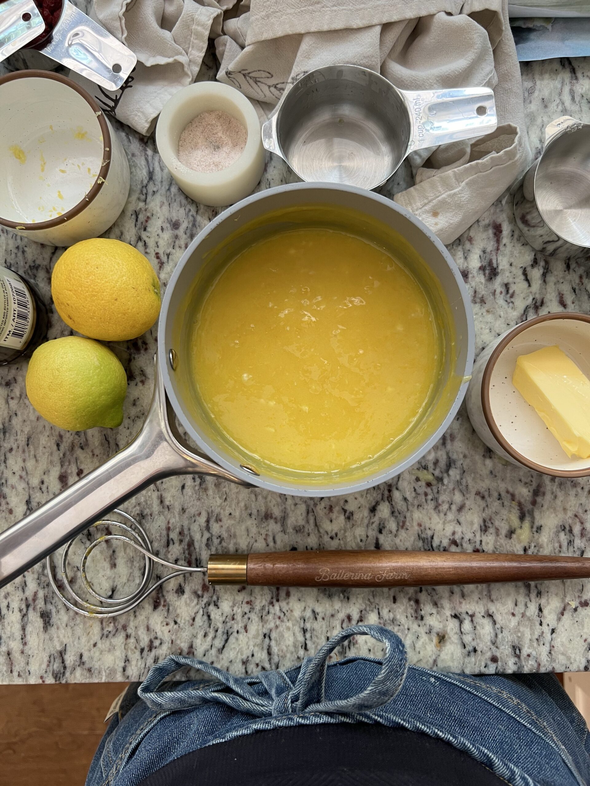 Aerial view of a kitchen countertop with a saucepan filled with yellow liquid, surrounded by lemons, butter, measuring cups, and a whisk.