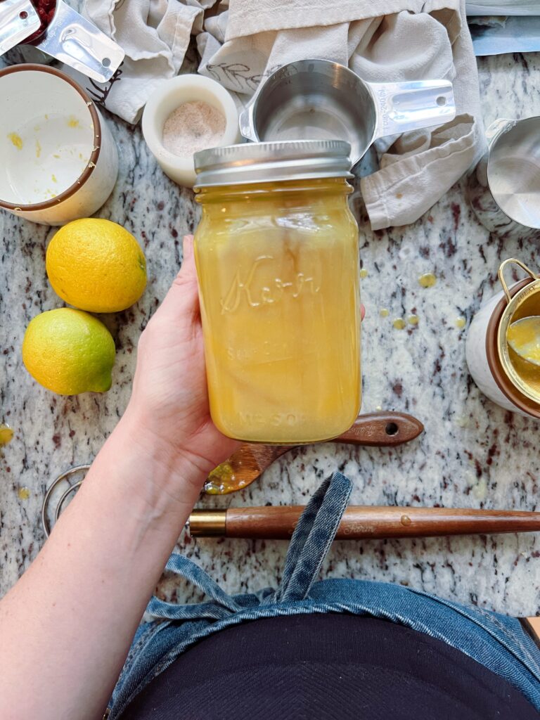 A person holding a jar filled with a yellow liquid, with lemons and baking utensils in the background on a marble countertop.