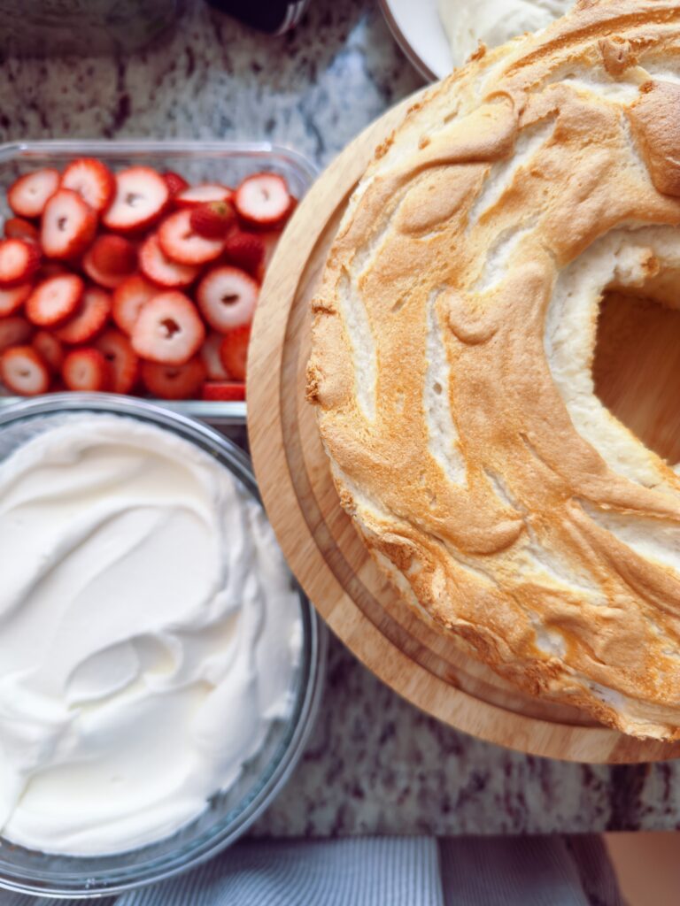A freshly baked angel food cake on a wooden cake stand, surrounded by sliced strawberries in a clear container and a bowl of whipped cream.