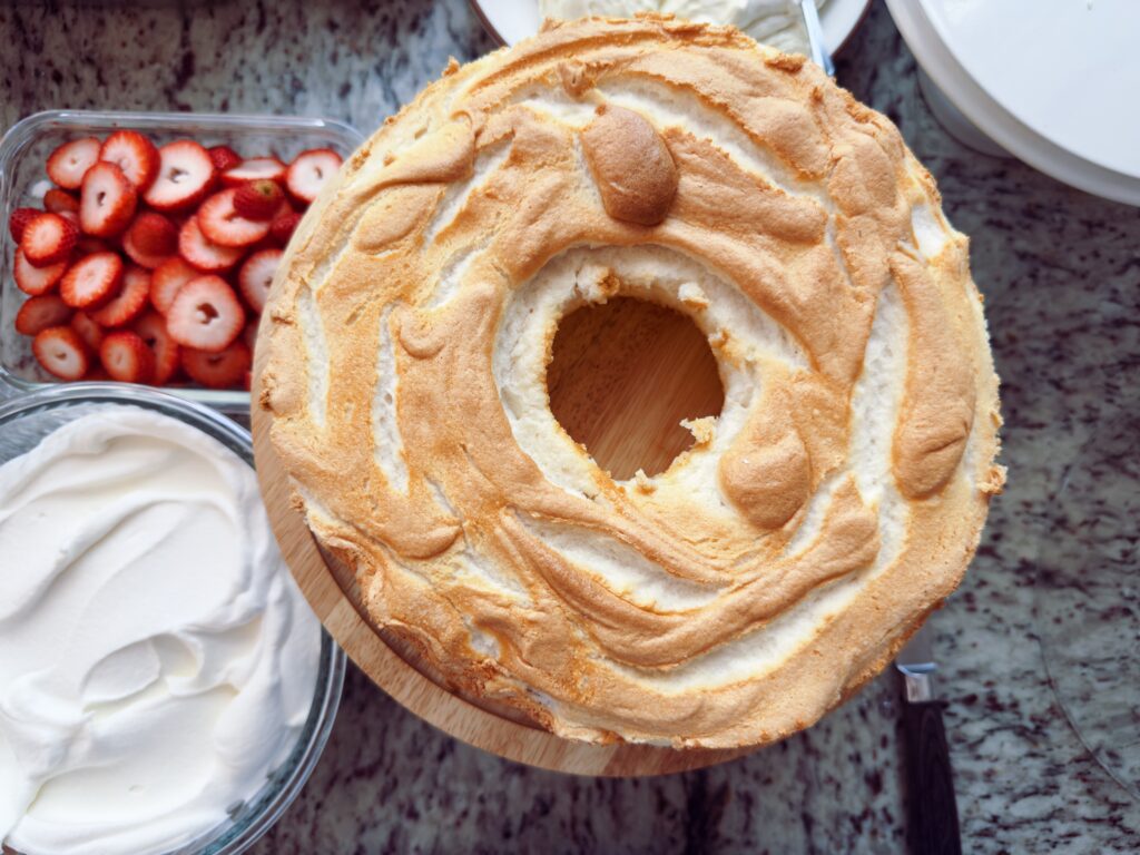 A top view of a fluffy angel food cake on a wooden serving board, surrounded by fresh sliced strawberries and a bowl of whipped cream.