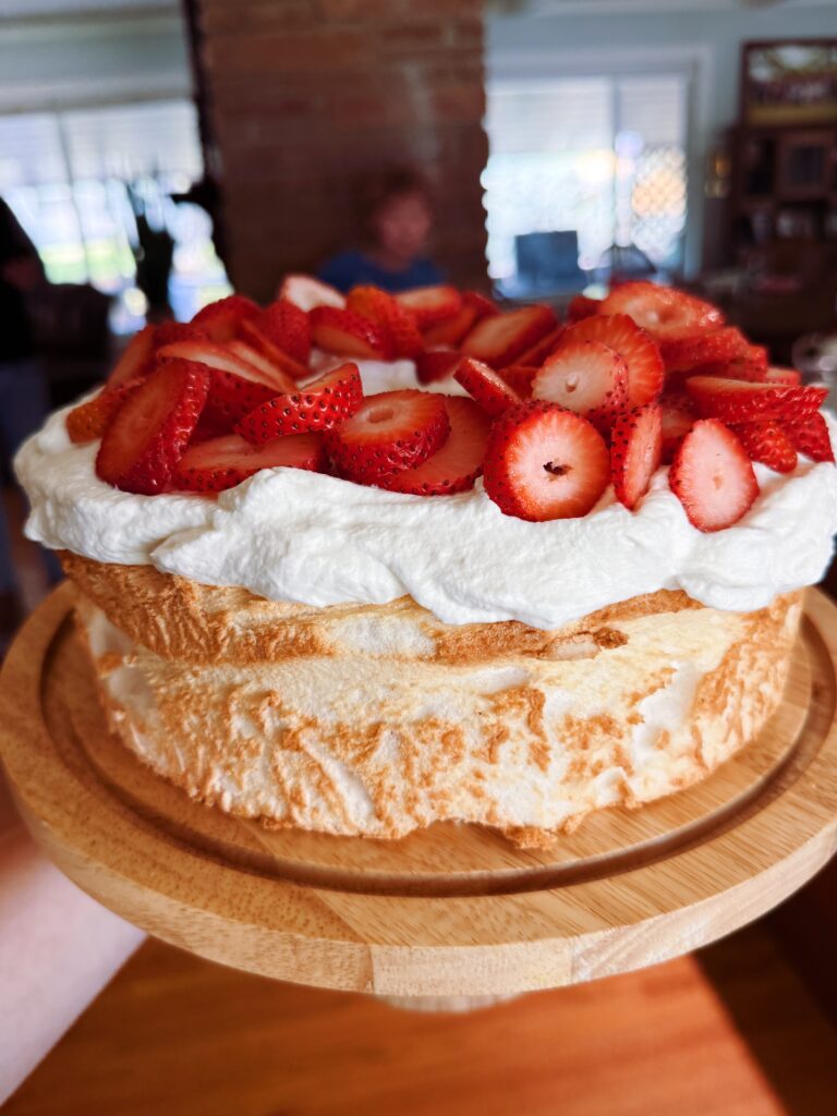 A freshly baked cake topped with whipped cream and sliced strawberries, presented on a wooden serving board.