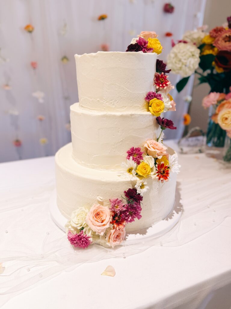 A three-tiered white wedding cake decorated with an assortment of colorful flowers cascading down one side.