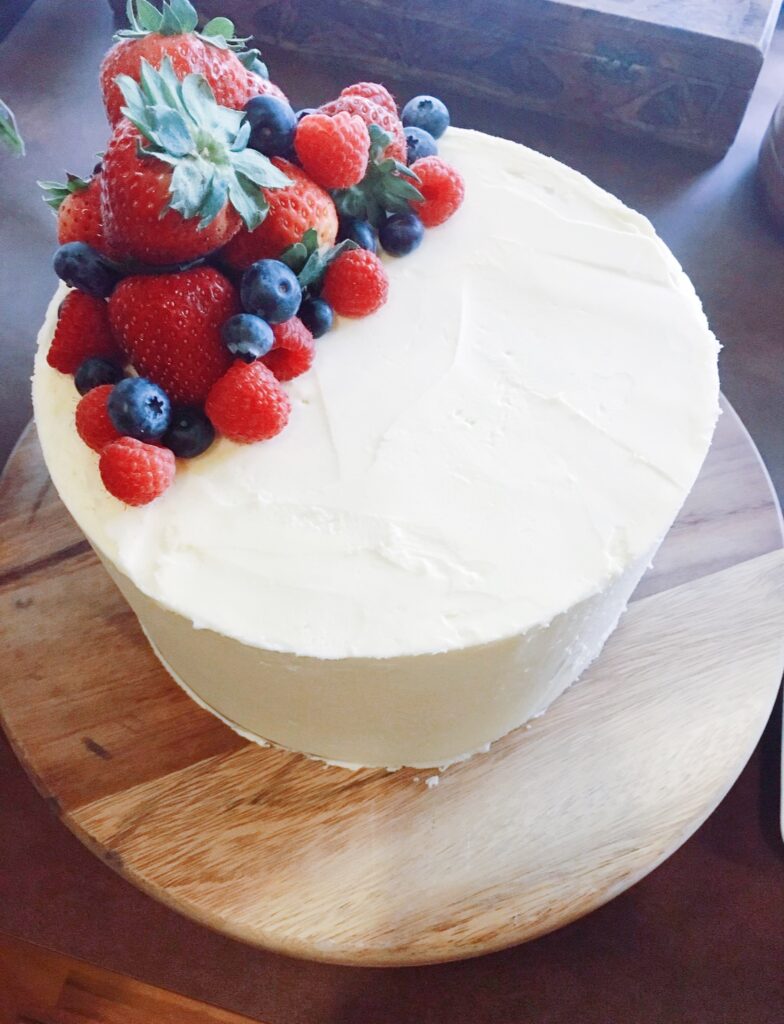A round cake with a smooth white frosting topped with a variety of fresh berries, including strawberries, raspberries, and blueberries, displayed on a wooden cake stand.