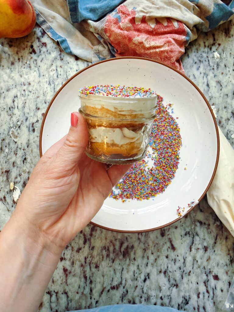 A hand holding a small glass jar filled with a layered dessert topped with colorful sprinkles, surrounded by scattered sprinkles on a plate and a messy kitchen countertop.