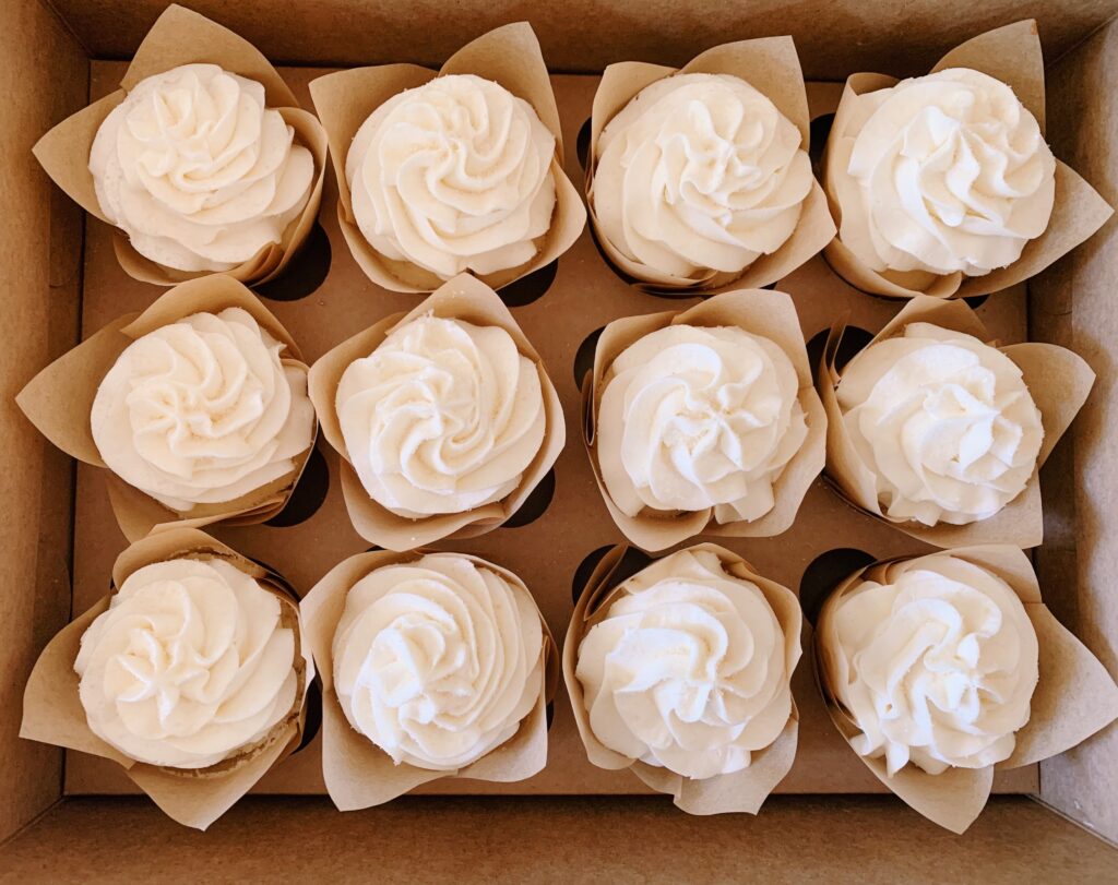 A box containing twelve cupcakes with swirled white frosting, arranged neatly in a grid.