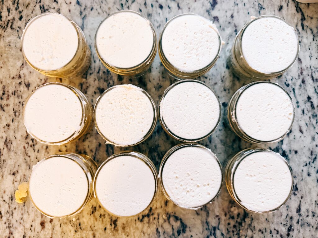 Twelve glass jars filled with a creamy white substance, arranged in four rows on a marble countertop.