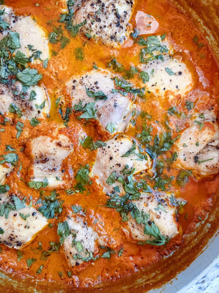 A close-up image of cooked chicken pieces in a rich orange sauce, garnished with fresh green herbs.