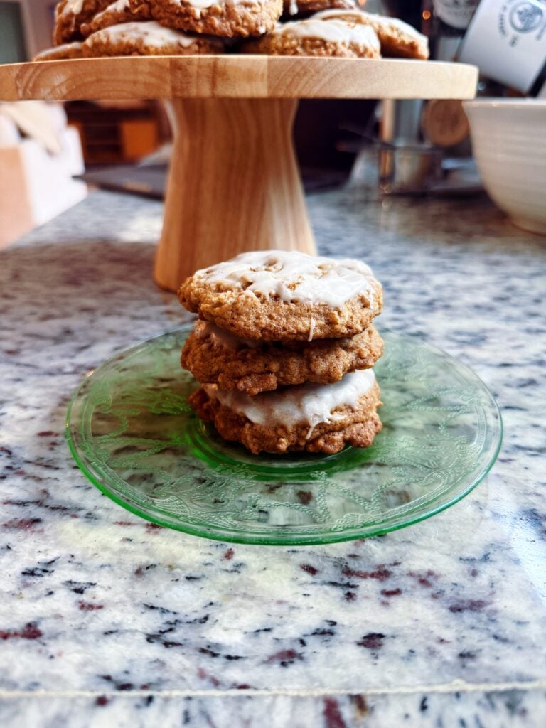 A stack of three oatmeal cookies with white icing on a green glass plate, placed on a marble countertop.